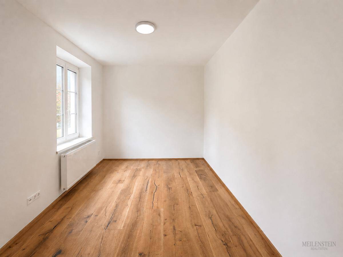 Leeres Zimmer mit hellem Holzboden und einem Fenster, das Tageslicht in den Raum bringt.