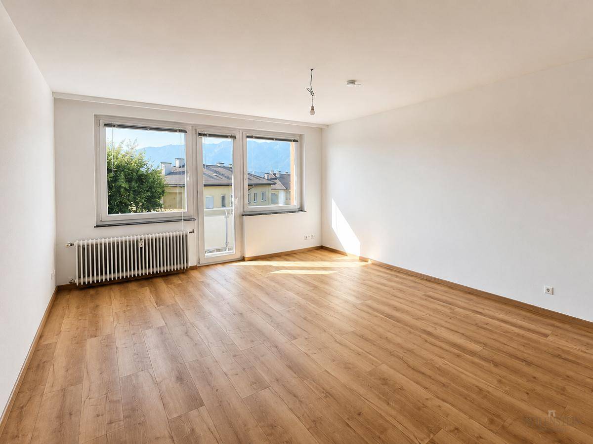 Heller Wohnraum mit Laminatboden, großen Fenstern und Zugang zu einem Balkon.