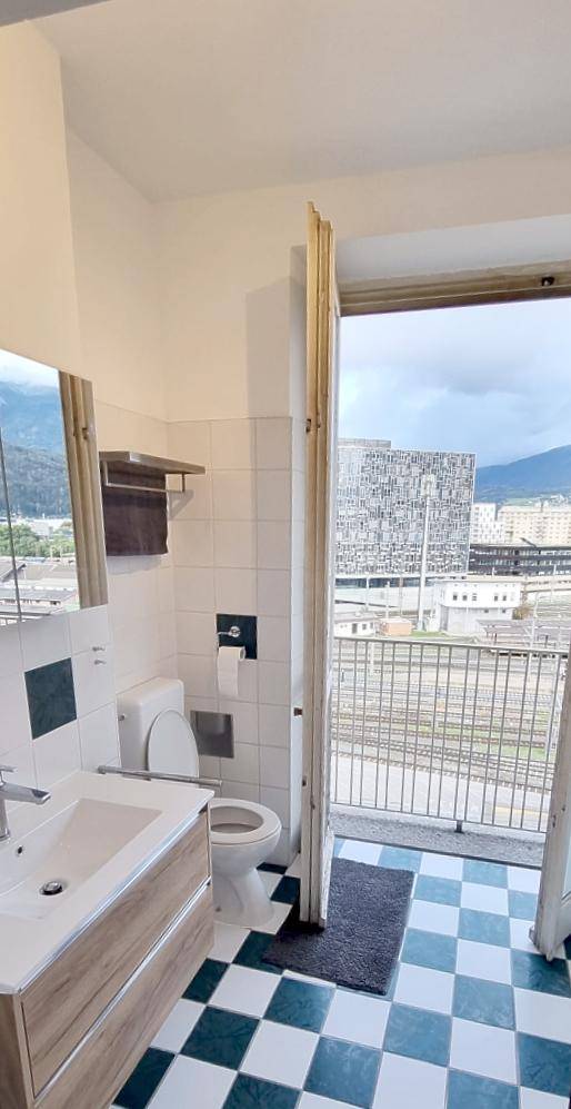 Badezimmer mit Balkonzugang, kariertem Fliesenboden und Blick auf die Stadt und Berge.