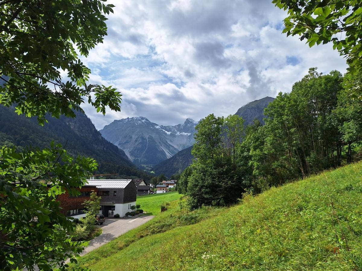 Weitläufiger Blick über ein grünes Tal mit Häusern und schneebedeckten Bergen unter bewölktem Himmel.