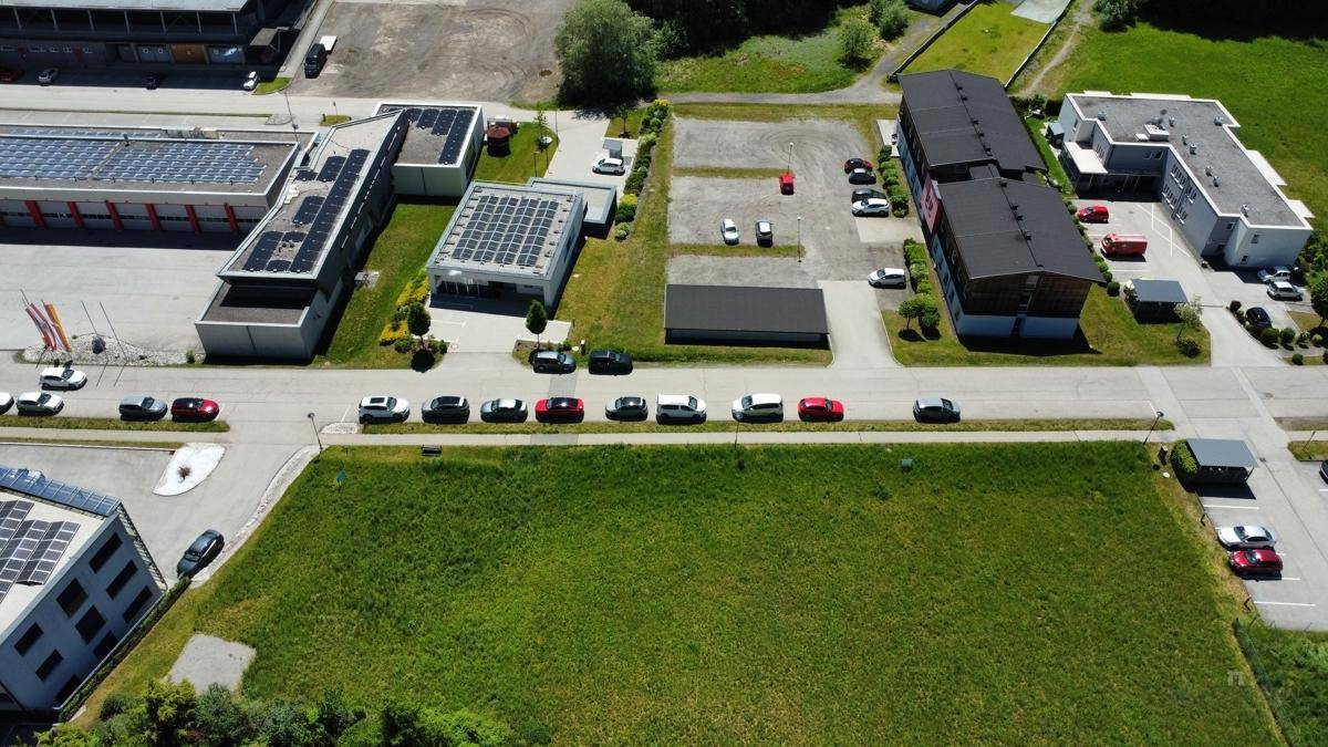 Luftaufnahme eines Gewerbegebiets mit mehreren Gebäuden, vielen geparkten Autos und einem großen grünen Grundstück im Vordergrund.