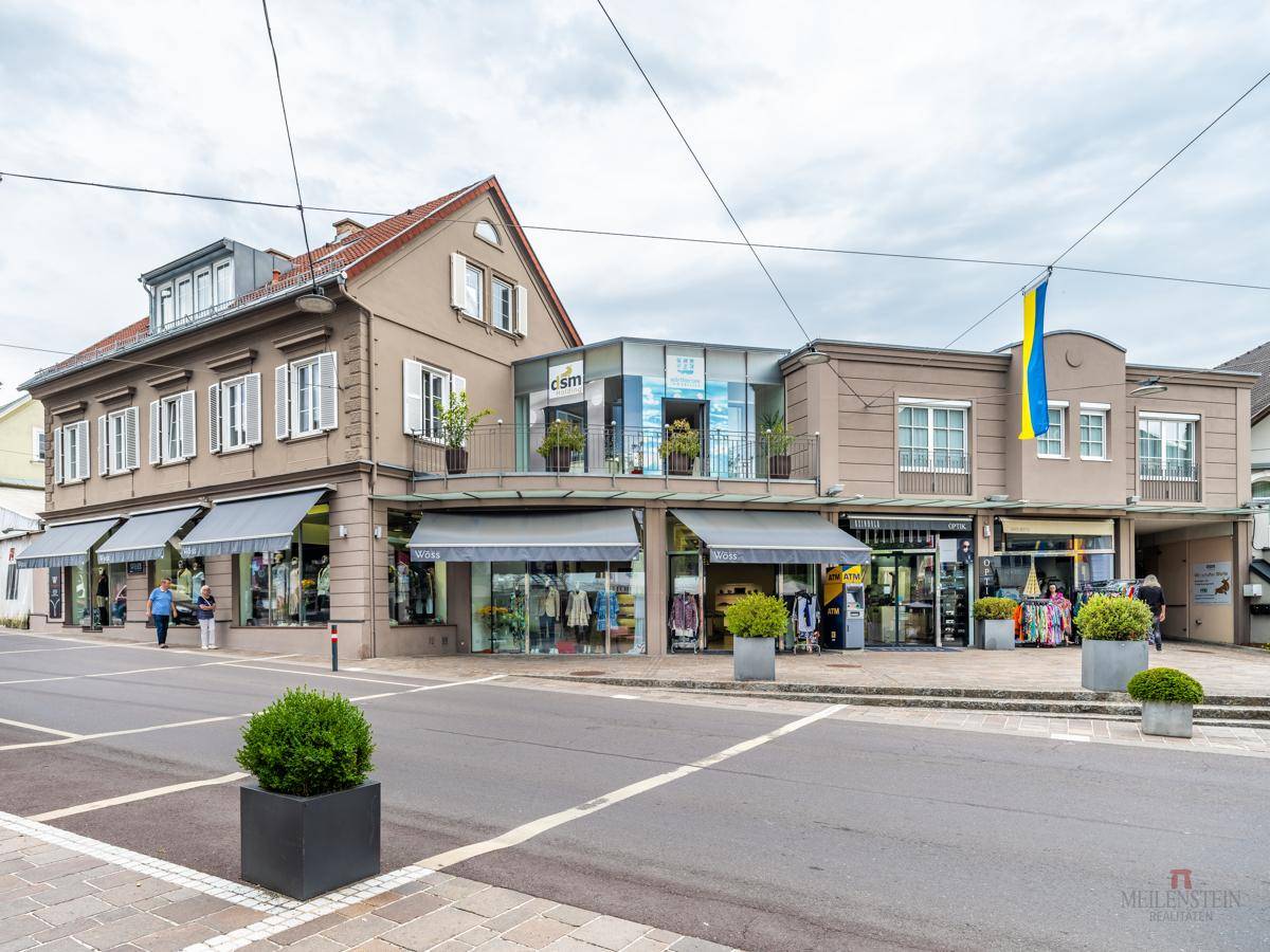 Tagesansicht einer Geschäftsstraße mit verschiedenen Geschäften, Markisen und einem modernen Gebäude mit Glasbalkon.