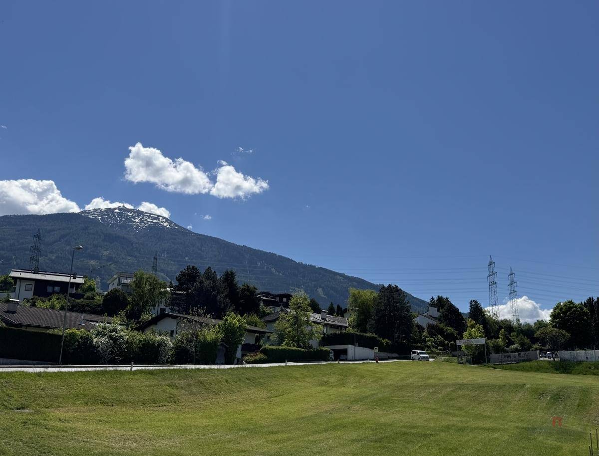Panoramablick über eine grüne Wiese mit Häusern und schneebedeckten Bergen im Hintergrund.
