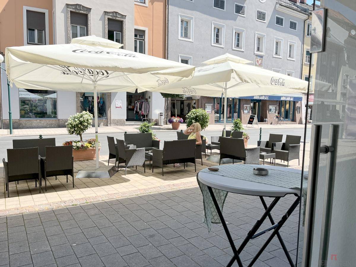 Belebter Außenbereich mit Sonnenschirmen und Tischen vor einem Gebäude in einer Stadtansicht.