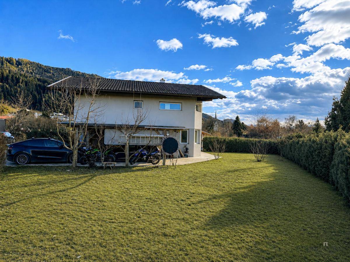 Seitenansicht des Hauses mit großem Rasenbereich, Bäumen und geparkten Fahrzeugen unter dem Gebäude.