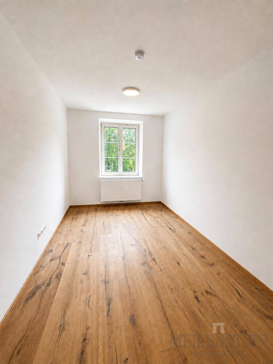 Geräumiges, leeres Zimmer mit Holzboden und einem Fenster, das eine angenehme Helligkeit schafft.