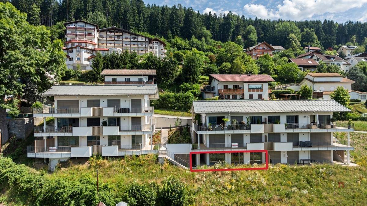 Zwei moderne Apartmentgebäude mit Balkonen am Hang, umgeben von üppigem Grün und einem roten Rahmen.