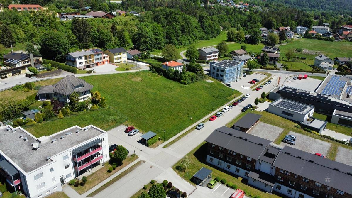 Überblick aus der Luft über ein großes grünes Baugrundstück, umgeben von Wohn- und Geschäftsgebäuden mit bewaldeten Hügeln im Hintergrund.