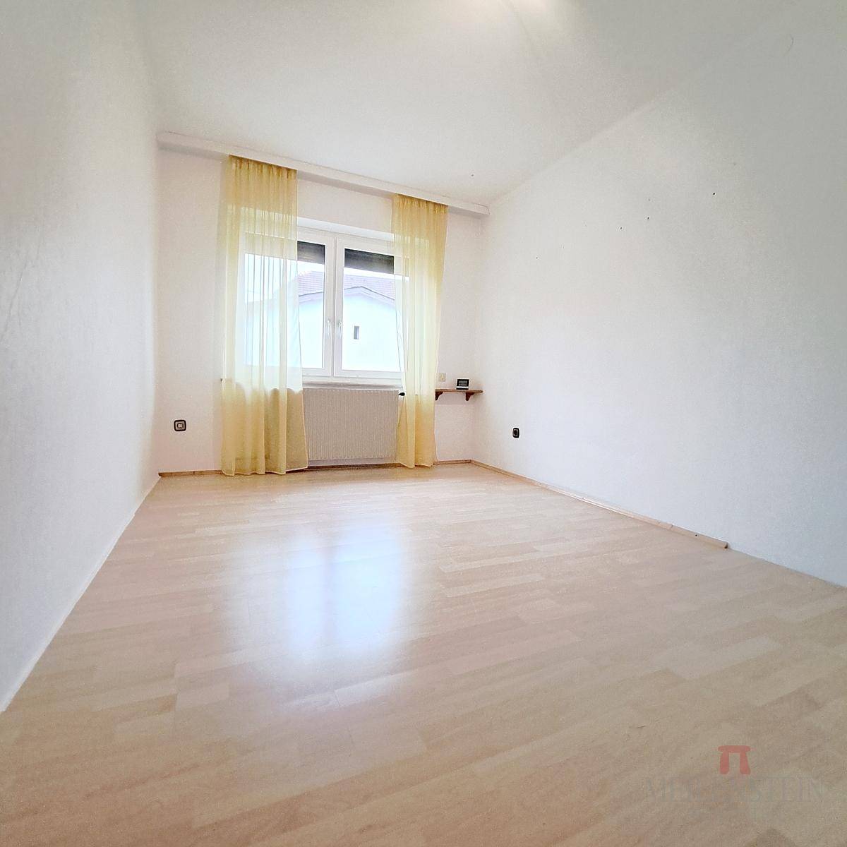 Helles, leeres Zimmer mit Laminatboden, weiß gestrichenen Wänden und großem Fenster.