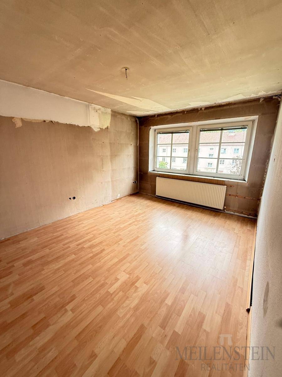 Helles Wohnzimmer mit Laminatboden und zwei Fenstern, das auf eine umfassende Renovierung wartet.