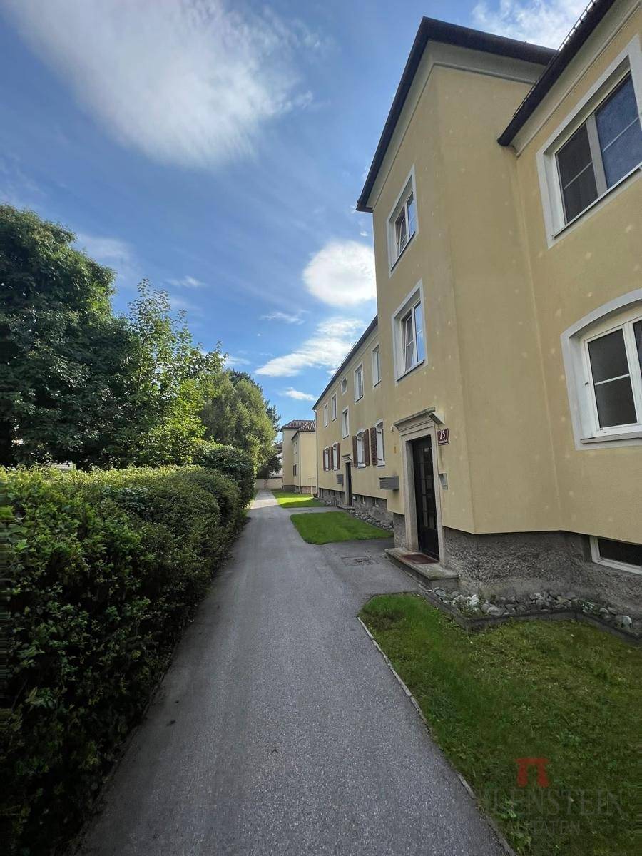 Gepflasterter Zugang zum Gebäude, gesäumt von einer hohen, grünen Hecke und Rasenflächen.