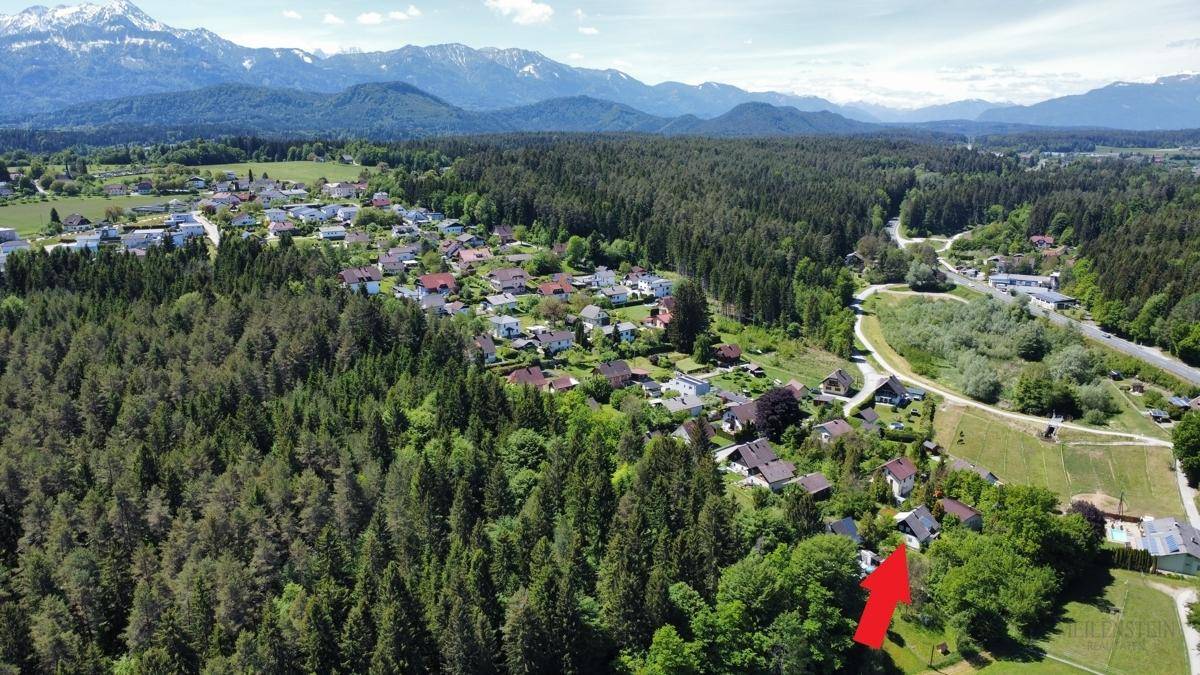 Luftaufnahme eines Wohngebiets umgeben von Wald und mit Bergblick, das Objekt ist mit einem roten Pfeil markiert.