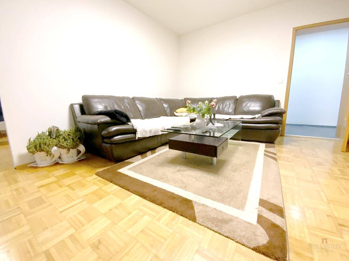 Geräumiges Wohnzimmer mit großem braunem Ledersofa, Glastisch und hellem Parkettboden.