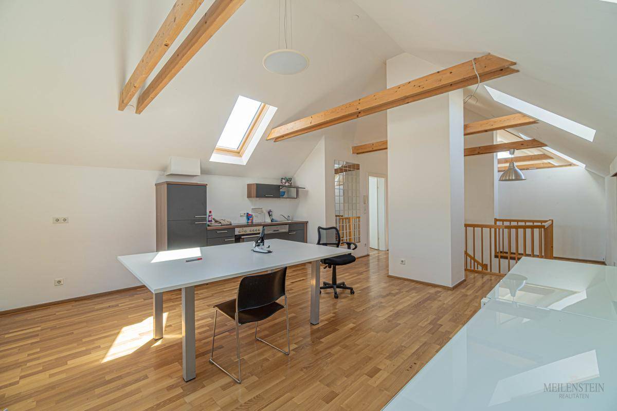 Heller Dachgeschossraum mit Holzboden, sichtbaren Holzbalken und Dachfenstern, inklusive Küchenzeile.