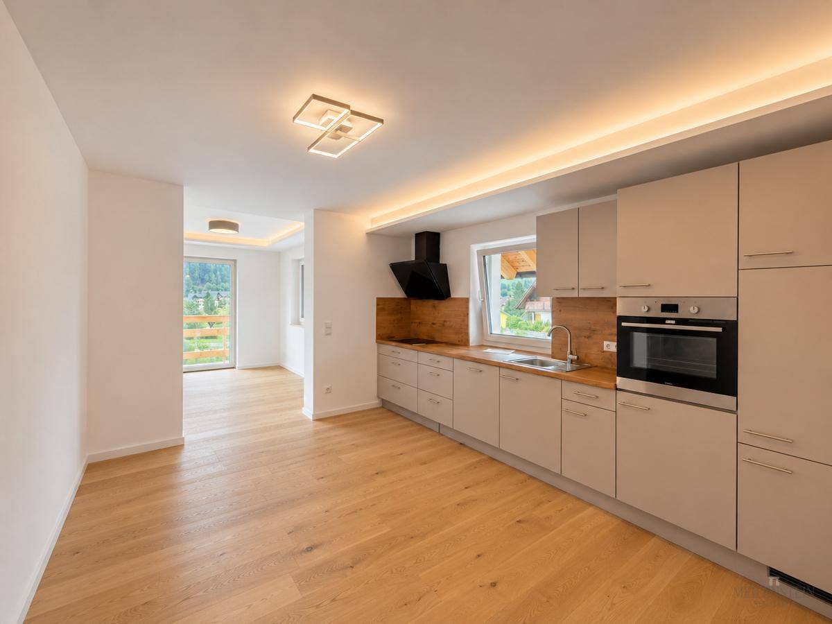 Moderne Einbauküche mit beigen Fronten und Holzboden, ausgestattet mit Backofen, offen zu einem hellen Wohnbereich.