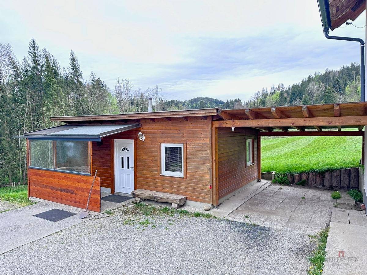 Holzgebäude mit Carport und einem kleinen Wintergarten, umgeben von grüner Landschaft.