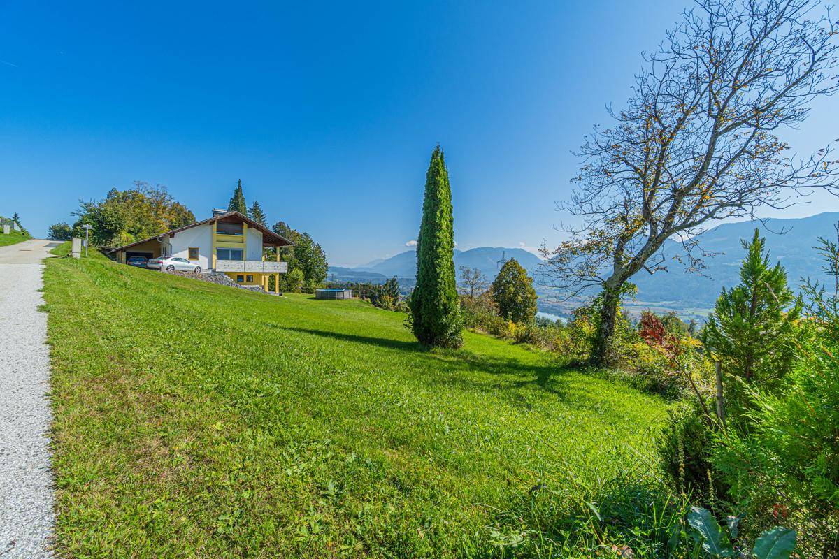Haus auf einem Hanggrundstück mit grüner Wiese, Zufahrt und malerischem Bergblick.