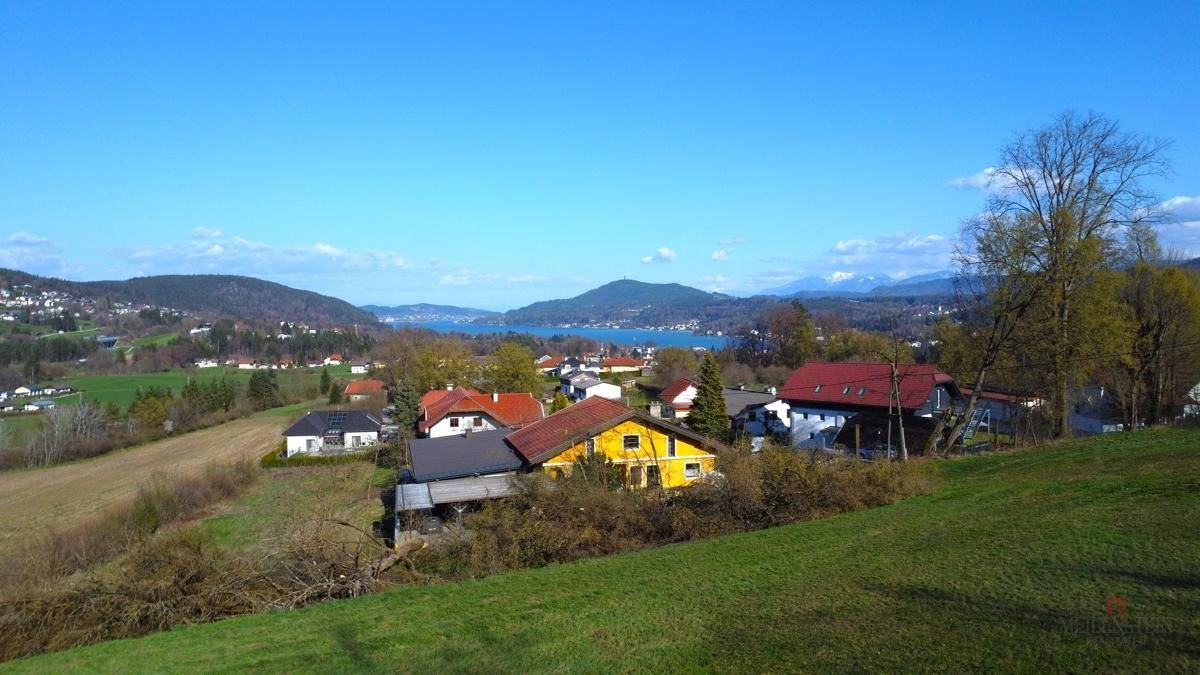 Blick über Wohnhäuser auf einem Hügel mit einem See und einer Bergkette am Horizont unter blauem Himmel.