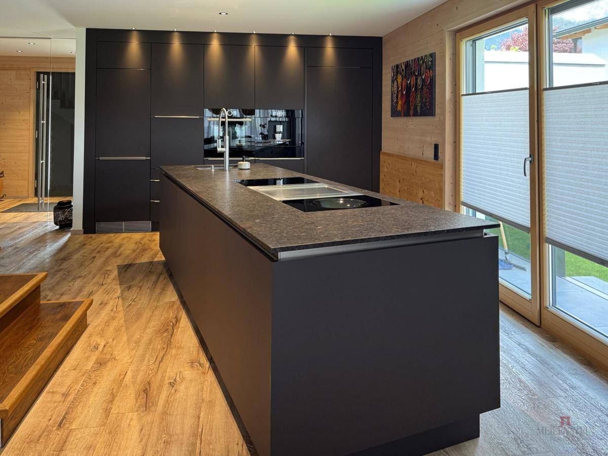 Moderne schwarze Küche mit Kochinsel, integrierten Geräten und Holzboden, hell beleuchtet.