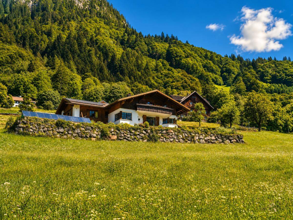 Traditionelles Haus in alpiner Landschaft mit grünen Wiesen und bewaldeten Bergen im Hintergrund.