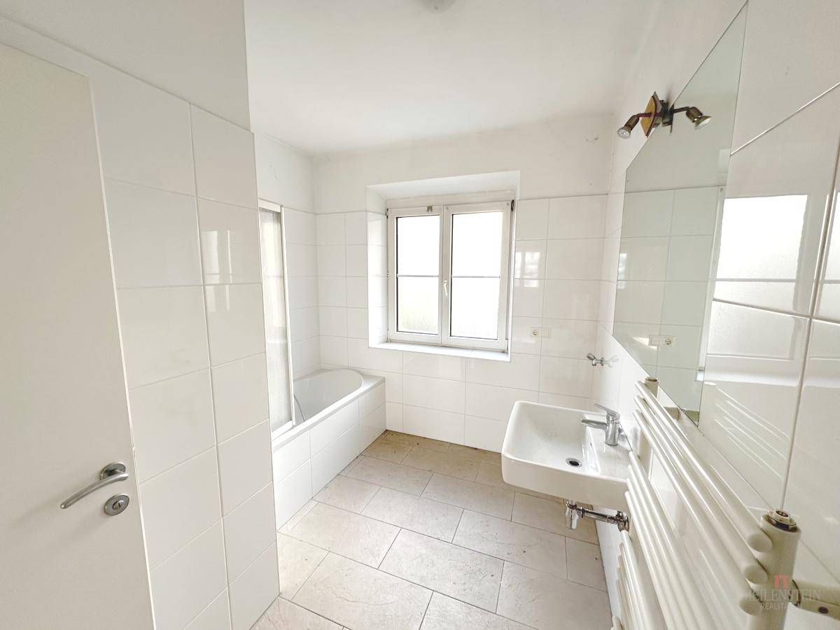Helles Badezimmer mit weißen Fliesen, einer Badewanne, Waschbecken und einem Fenster.