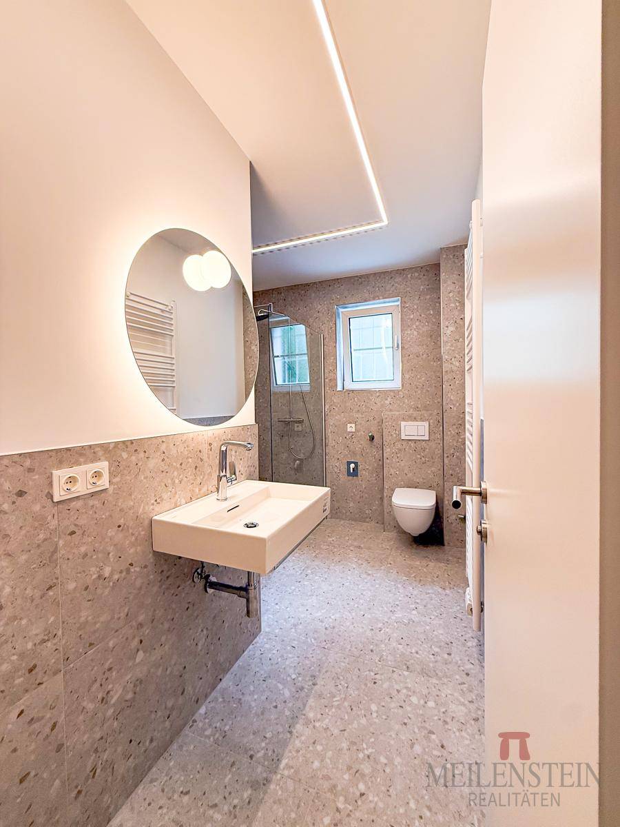 Modernes Badezimmer mit hellen Fliesen, wandhängendem Waschbecken, WC und beleuchtetem Spiegel.