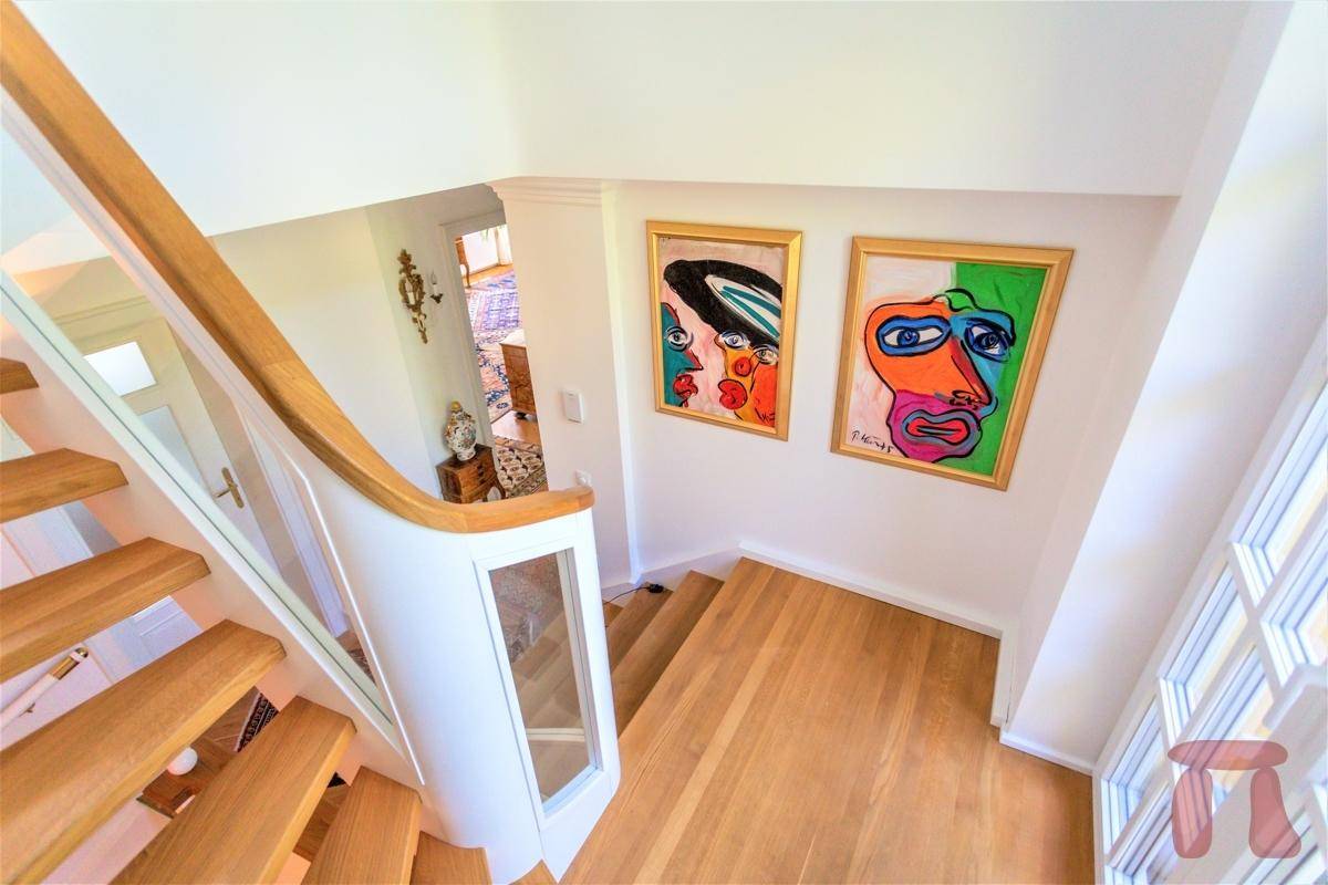 Helles Treppenhaus mit Holzstufen, Glasgeländer und farbenfrohen Kunstwerken.