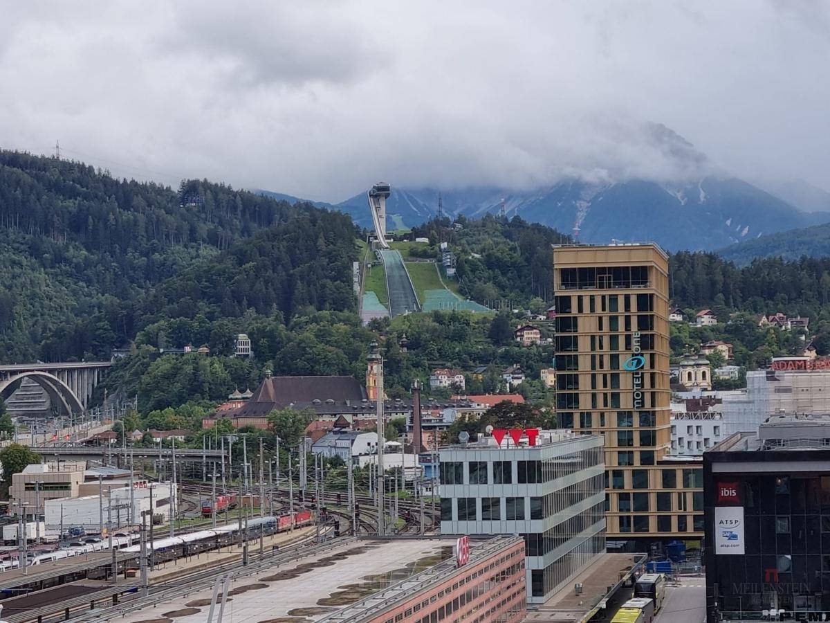 Stadtansicht mit Skisprungschanze, grünen Hügeln und Bergen unter bewölktem Himmel.