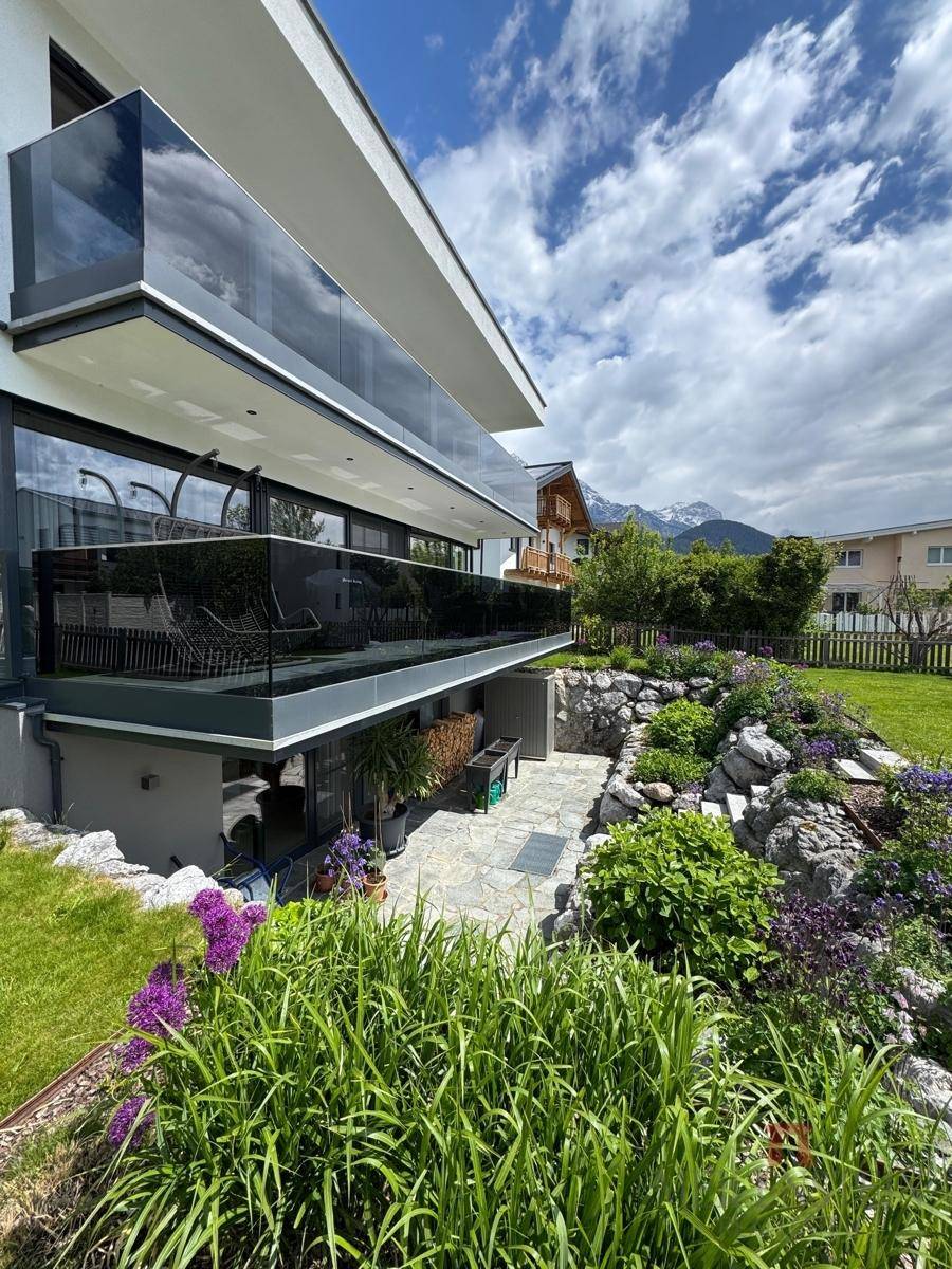 Seitliche Ansicht des modernen Gebäudes mit Terrasse und blühendem Gartenbereich vor Bergkulisse.