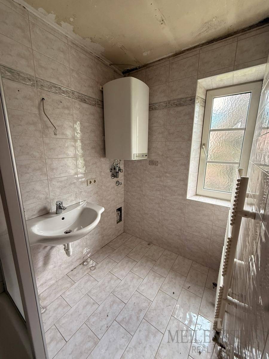 Gefliestes Badezimmer mit Waschbecken, Warmwasserboiler und Fenster, das eine Modernisierung erfordert.