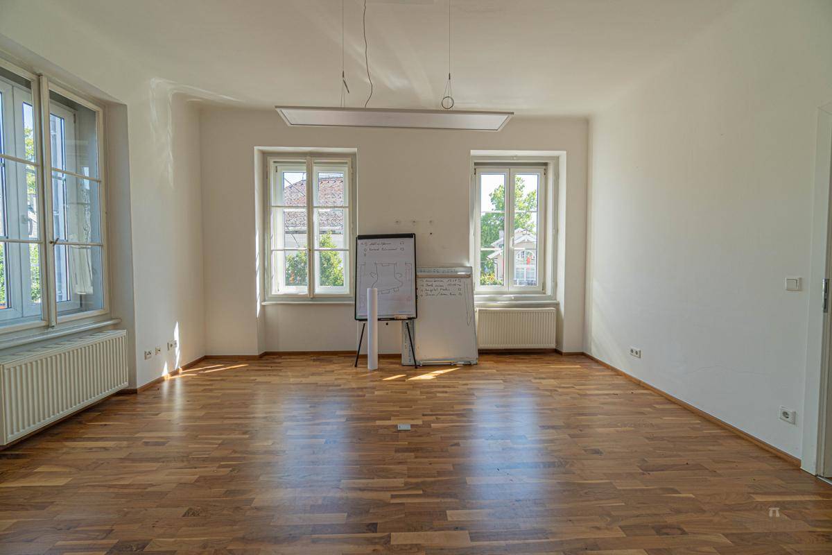 Heller Büroraum mit Holzboden und zwei großen Fenstern, die viel natürliches Licht spenden.