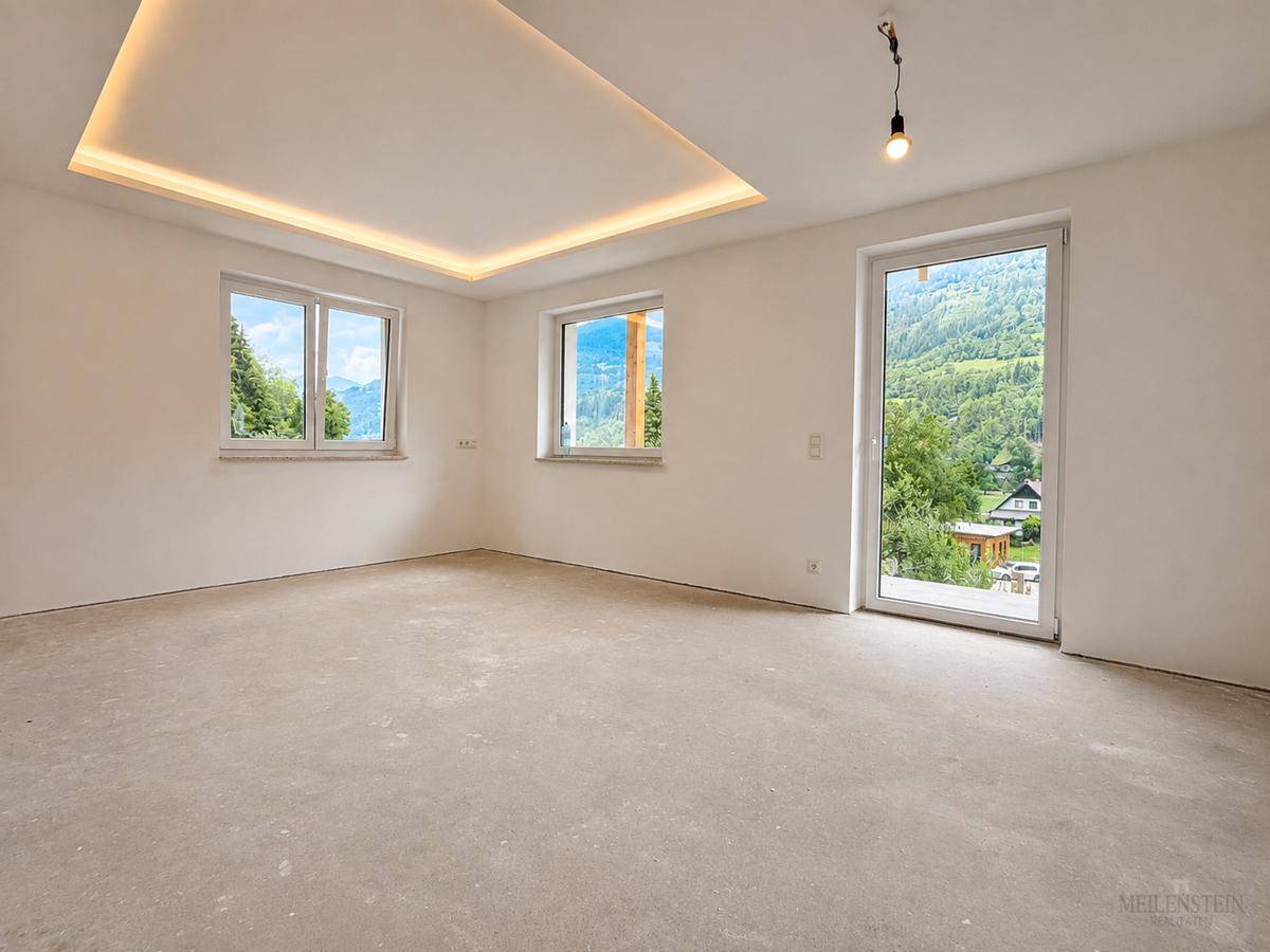 Geräumiger, unfertiger Raum mit Betonboden, großen Fenstern und Bergblick, ideal für einen Wohnbereich.