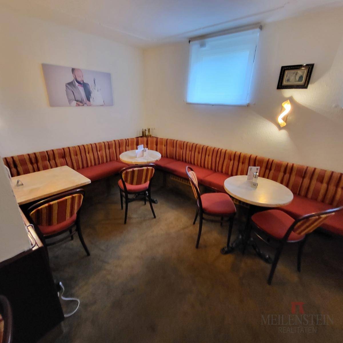 Eckbereich eines Cafés mit langer, gepolsterter Sitzbank, kleinen Tischen und einem Fenster.