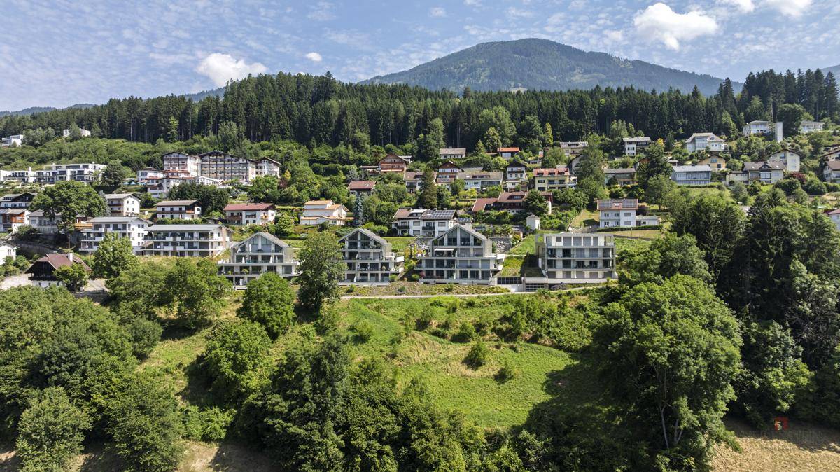Reihe moderner Wohngebäude am Hang, eingebettet in grüne Natur mit bewaldeten Bergen im Hintergrund.