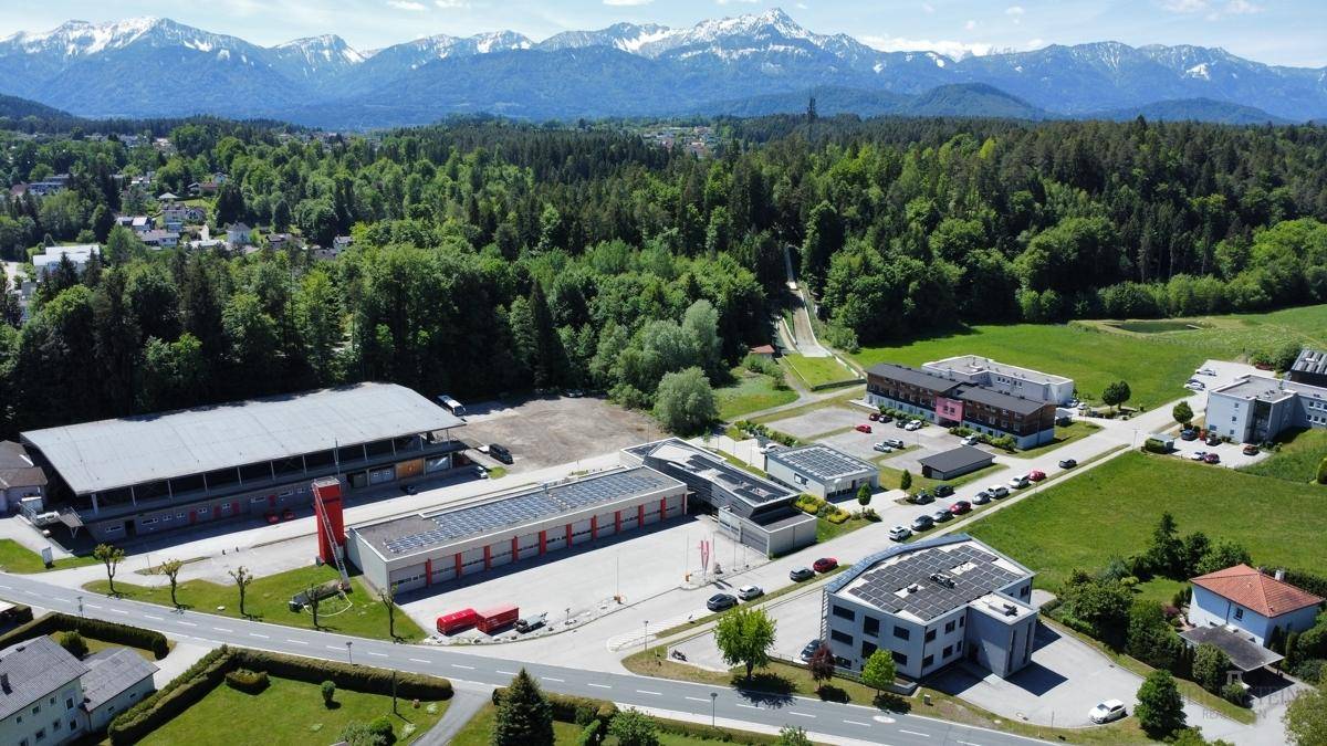 Panoramablick aus der Luft auf eine ländliche Gegend mit Gewerbegebäuden, weiten Grünflächen und einer beeindruckenden Bergkulisse.