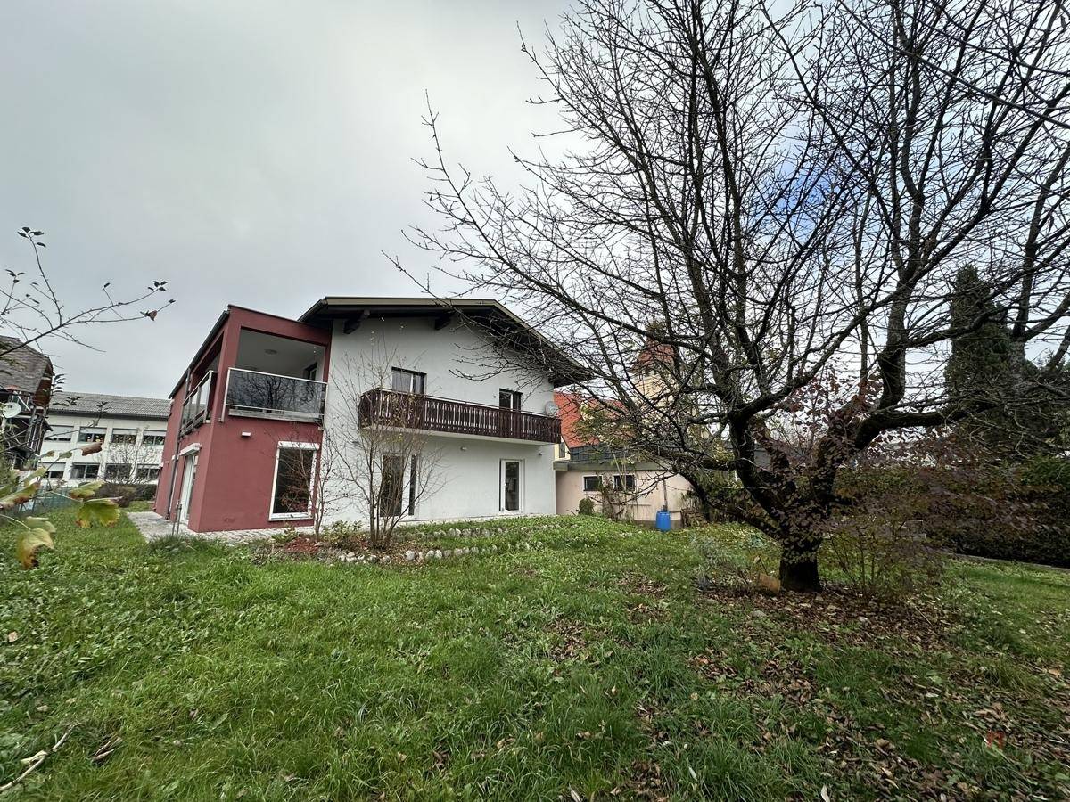 Einfamilienhaus mit weißer Fassade und rotem Anbau, umgeben von einem Garten mit altem Baum.