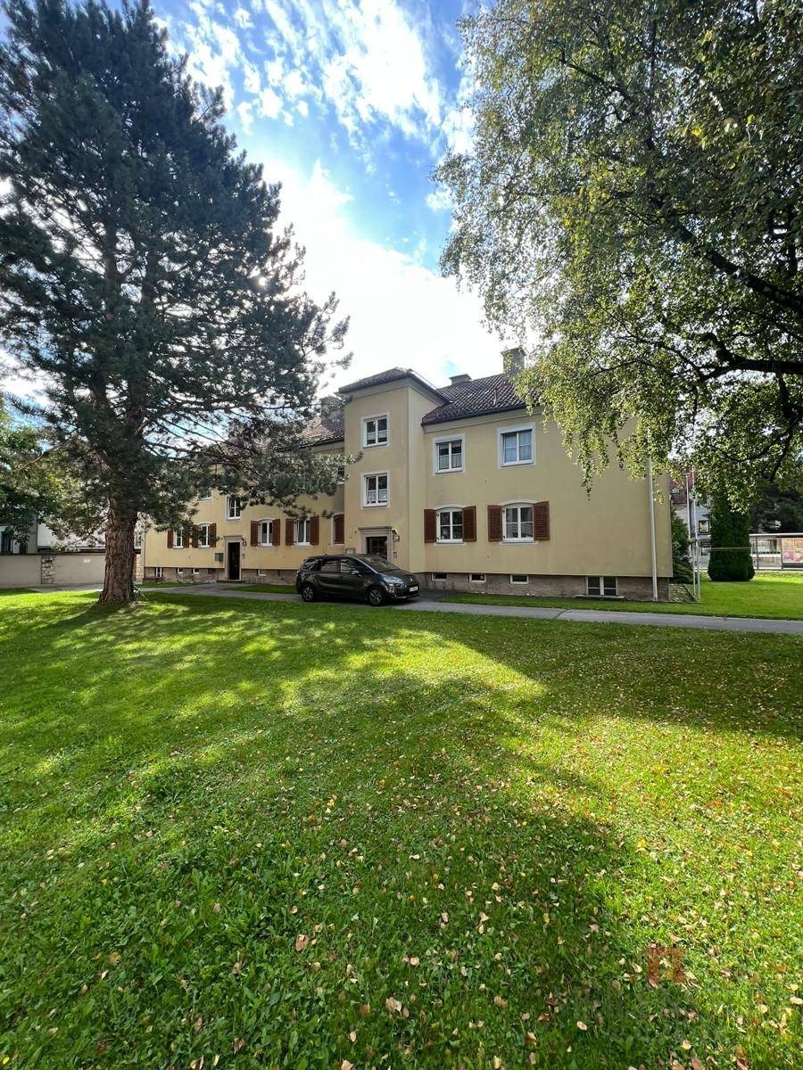 Das Mehrfamilienhaus mit seiner gelben Fassade und dem grünen Vorgarten unter strahlendem Himmel.