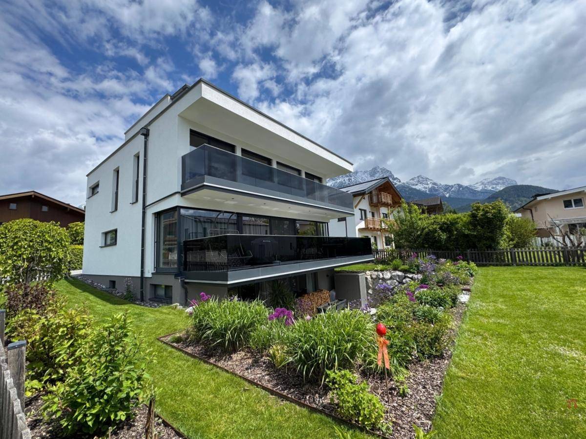 Elegantes modernes Gebäude mit Balkonen und einem schön angelegten Garten vor Bergpanorama.