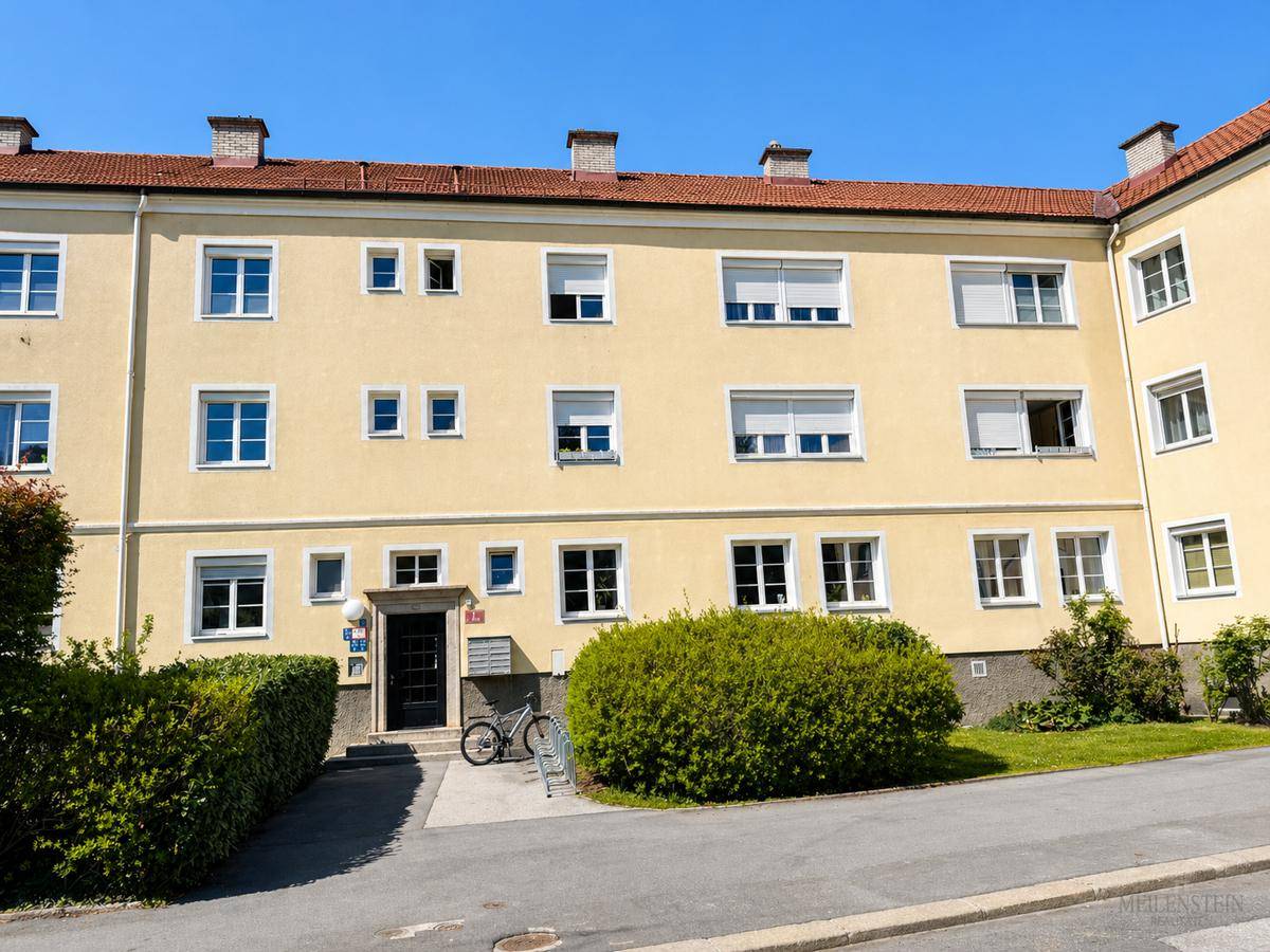 Wohngebäude mit heller Fassade, Eingangsbereich und Fenstern mit Rollläden, umgeben von Büschen.