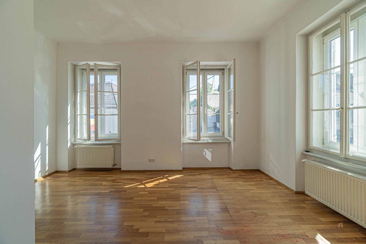 Geräumiger Raum mit Holzboden und drei großen Fenstern, die für eine helle und offene Atmosphäre sorgen.