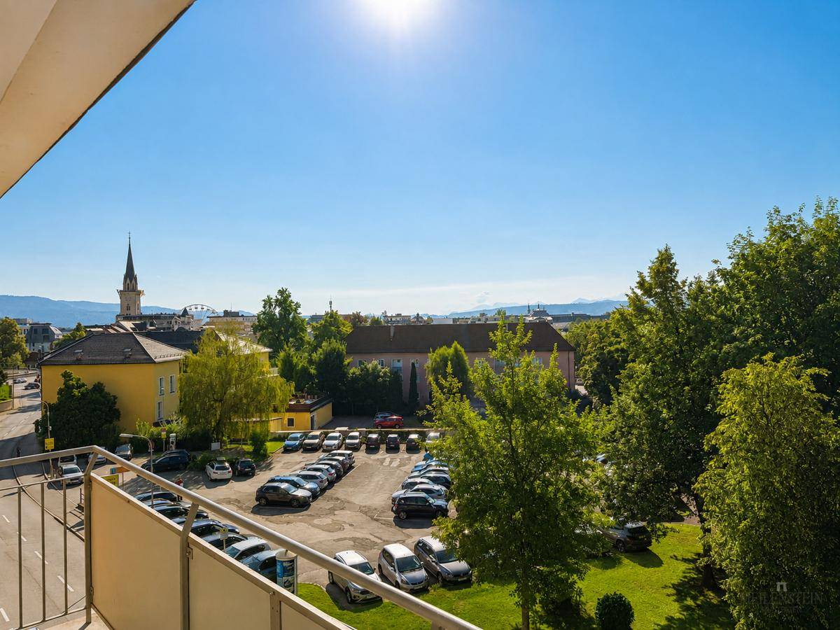 Sonniger Balkon mit Blick auf einen Parkplatz, städtische Gebäude und die umliegende Berglandschaft.