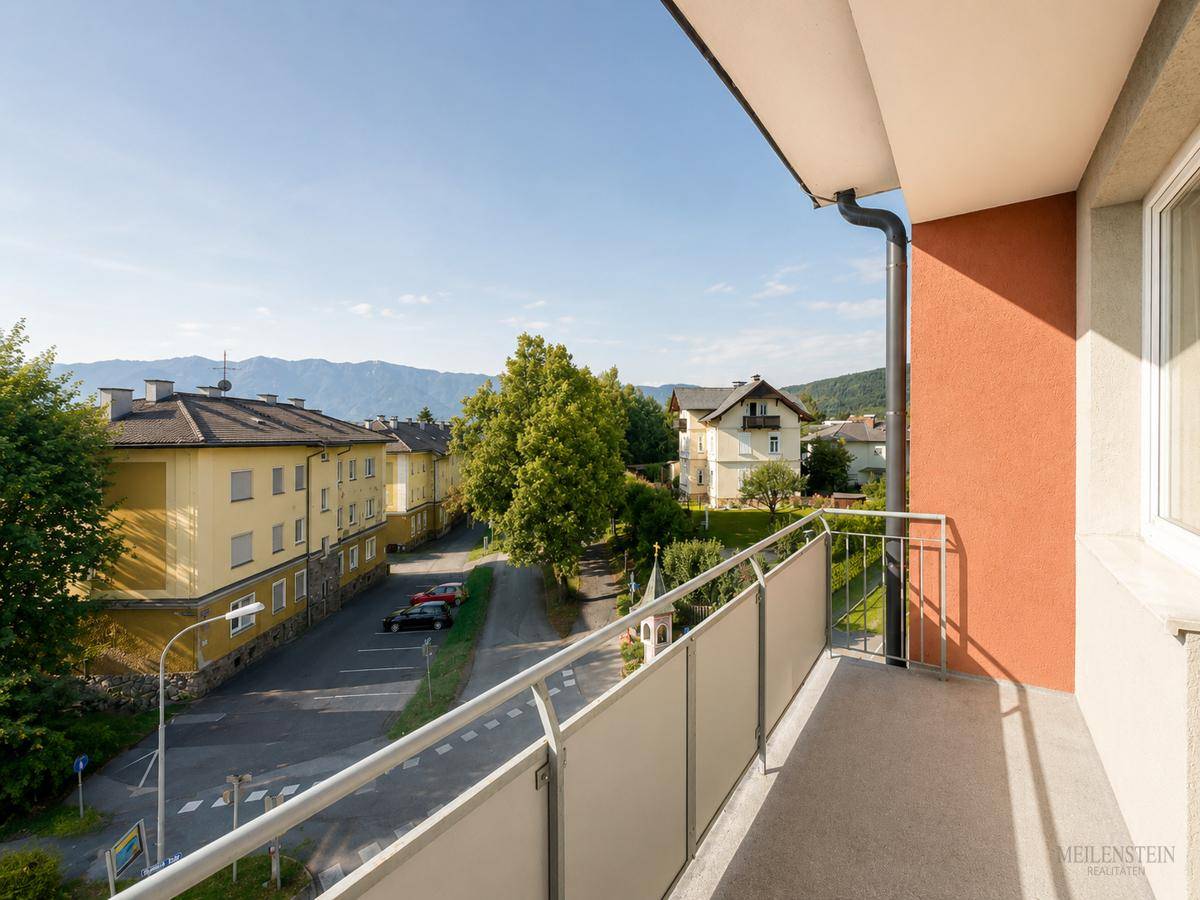 Balkon mit Blick auf eine ruhige Straße, weitere Gebäude und die bewaldeten Berge im Hintergrund.