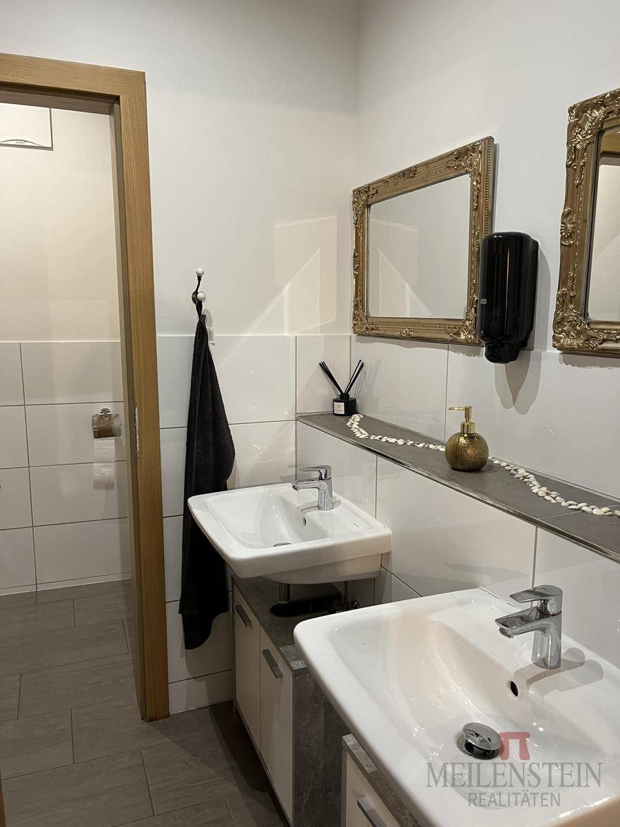 Badezimmer mit zwei Waschbecken, weißen Fliesen und zwei verzierten goldenen Spiegeln.