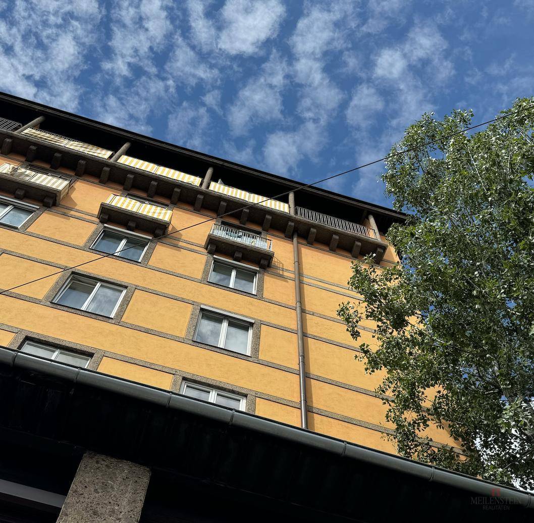 Fassade eines Gebäudes mit mehreren Balkonen und einem Baum im Vordergrund unter blauem Himmel.