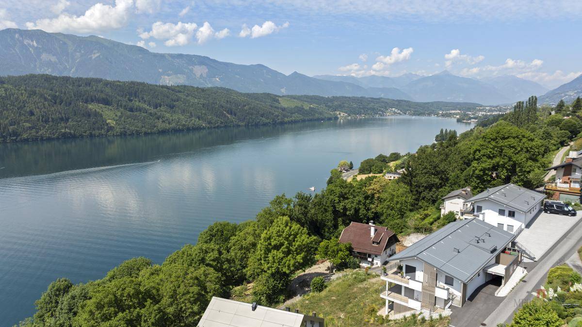 Weitläufige Ansicht des Sees mit modernen Häusern am Hang und majestätischen Bergen im Hintergrund.