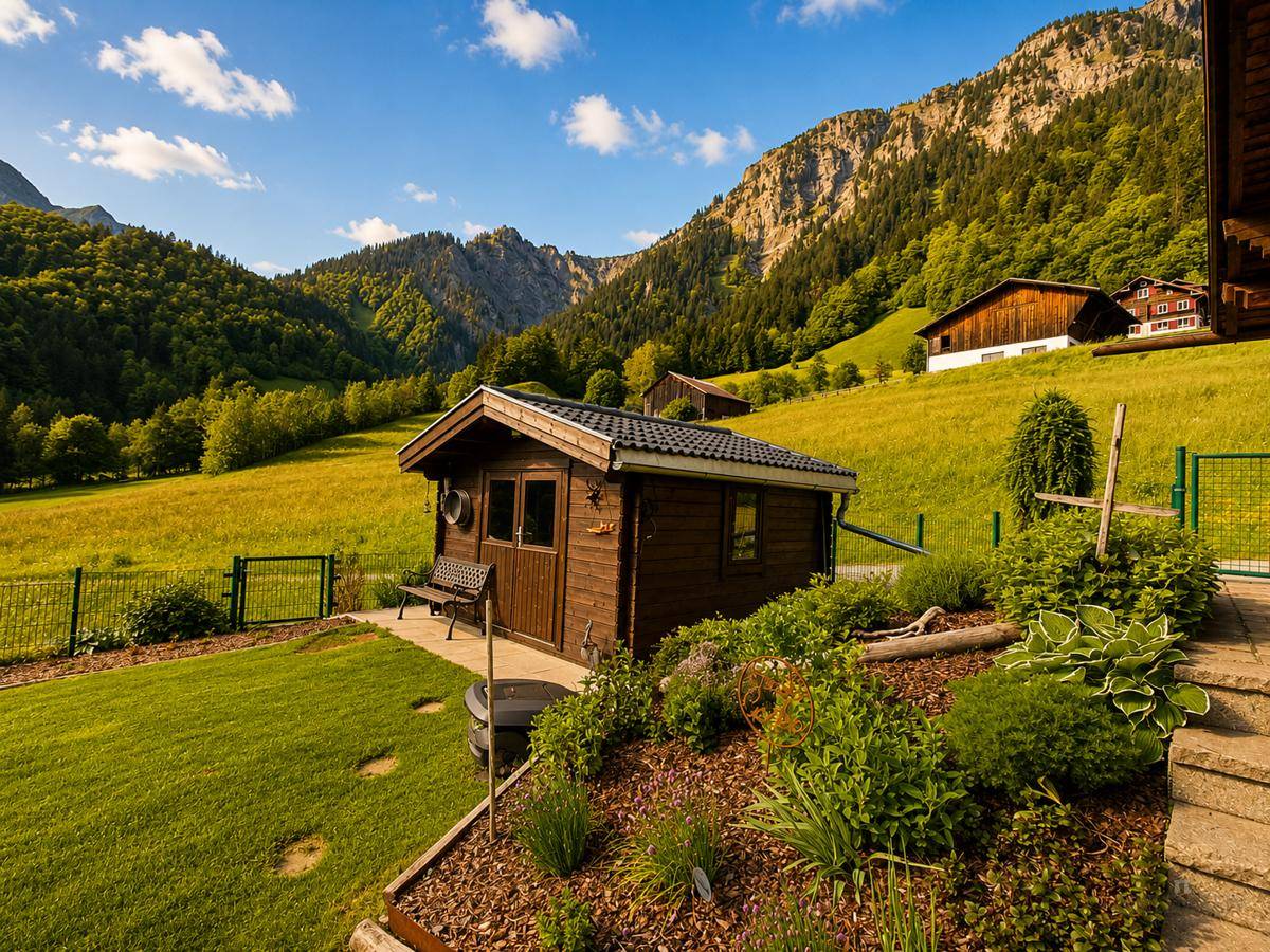 Kleines Holzgartenhaus in einer grünen Wiese, umgeben von Bergen unter blauem Himmel.
