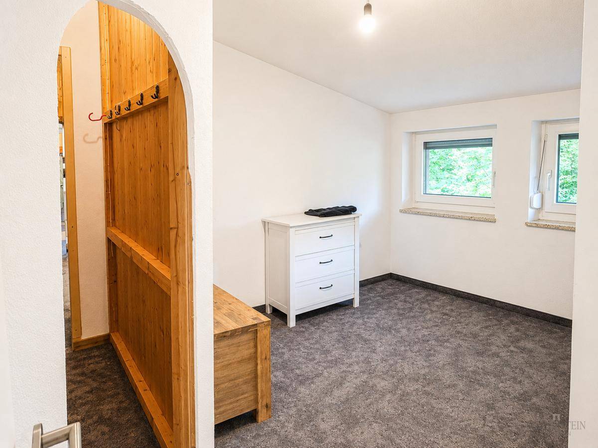 Helles Schlafzimmer mit Teppichboden, weißer Kommode und Fenstern mit Blick ins Grüne.