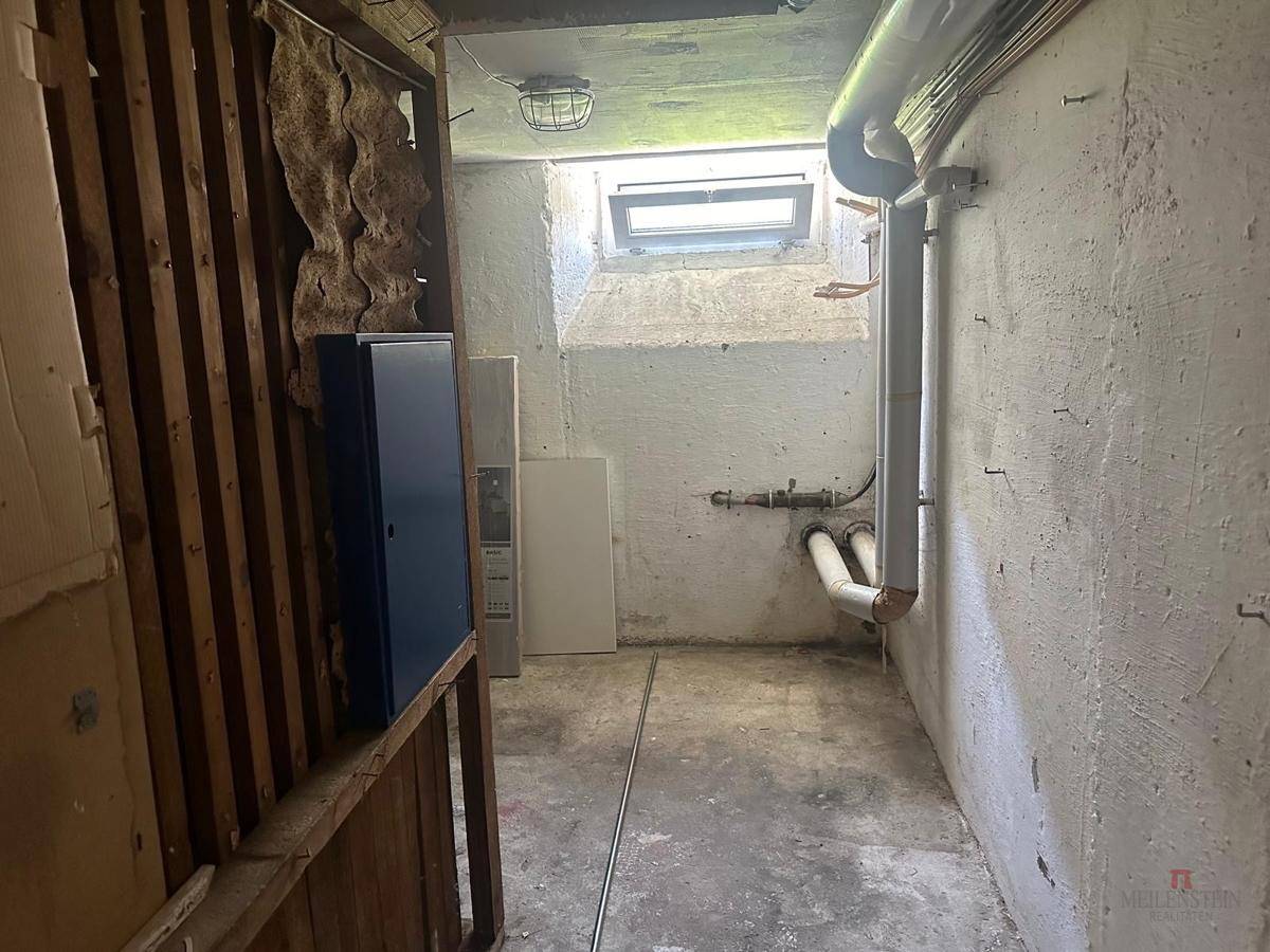 Abstellraum im Keller mit sichtbaren Rohren und einem kleinen Fenster, renovierungsbedürftig.