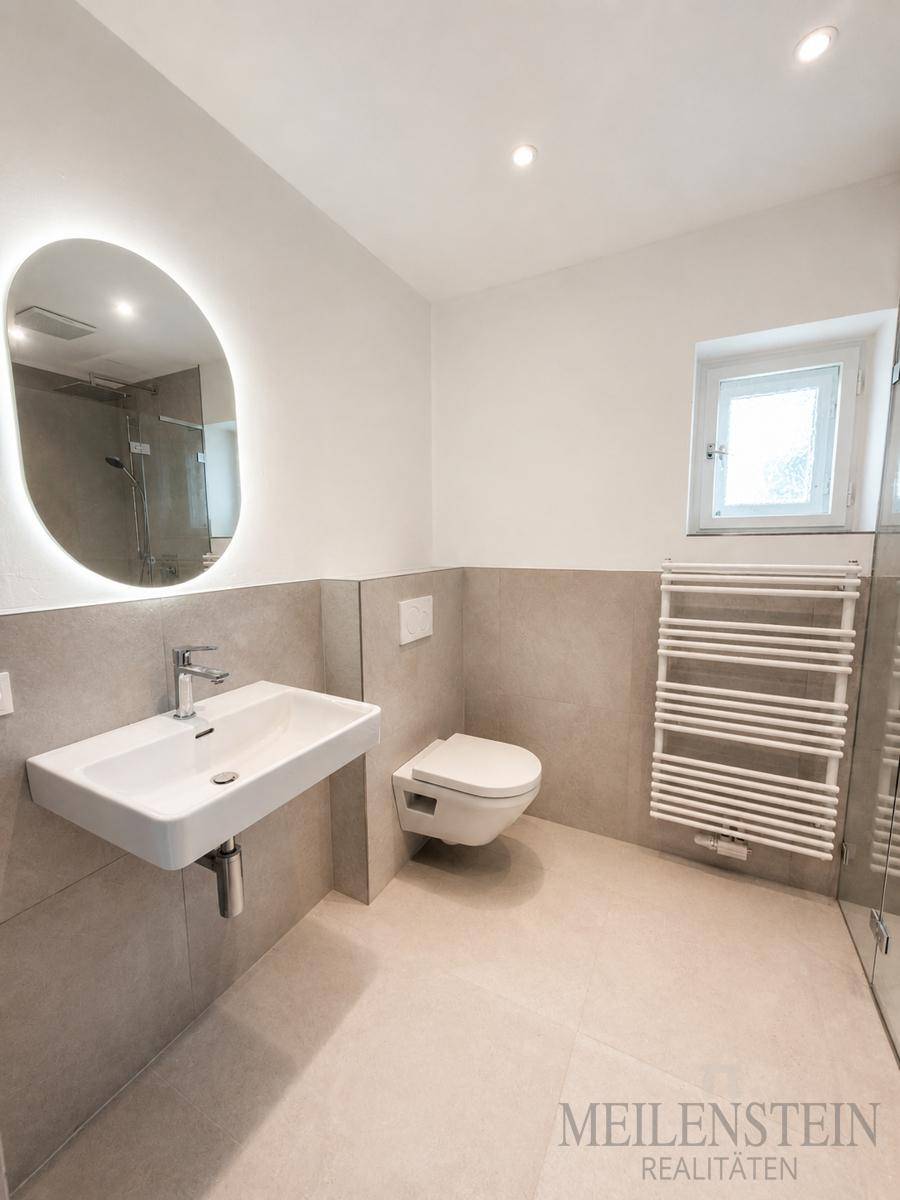 Modernes Badezimmer mit beleuchtetem Spiegel, Waschbecken und Wand-WC, in hellen Grautönen gehalten.