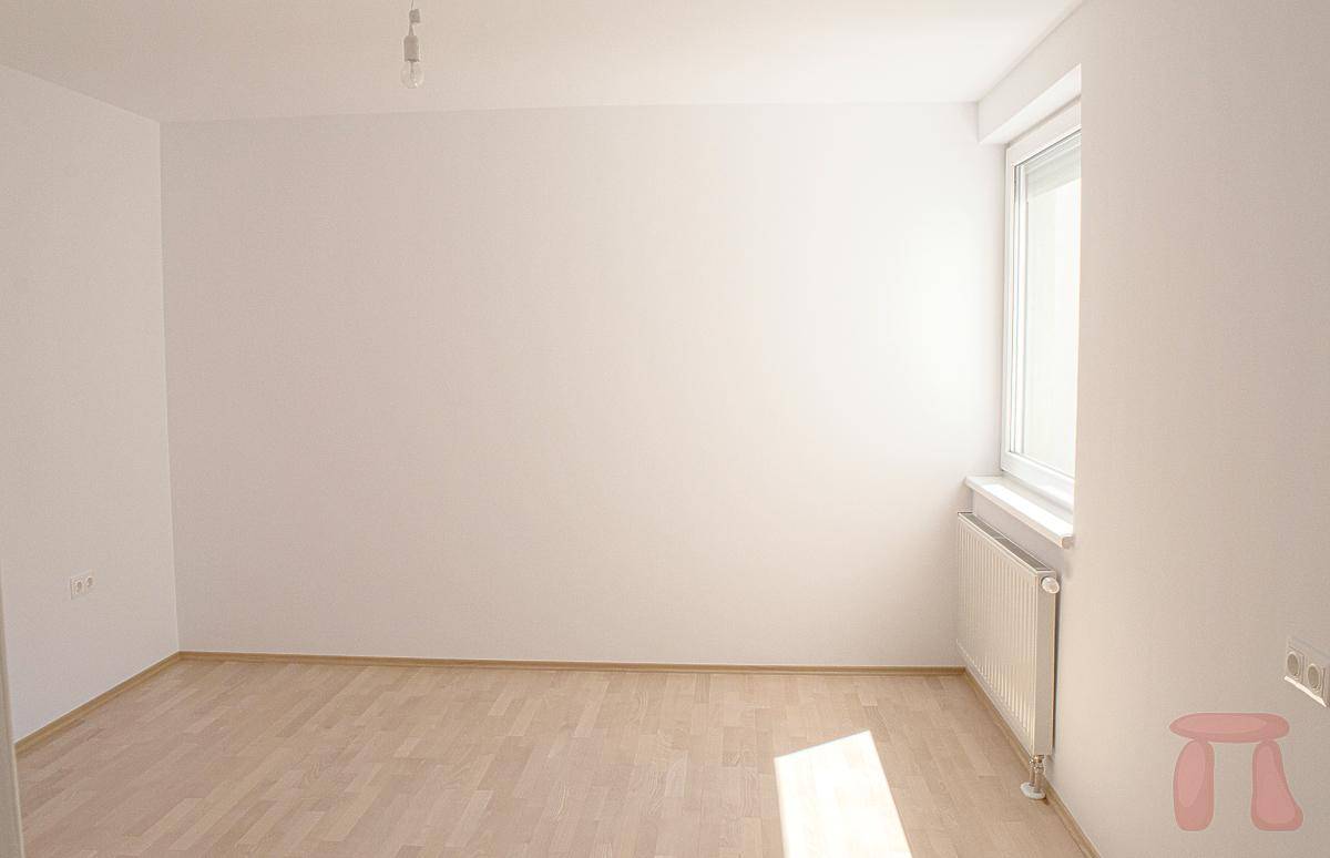 Leeres Schlafzimmer mit hellem Parkettboden und einem Fenster, das Sonnenlicht in den Raum lässt.